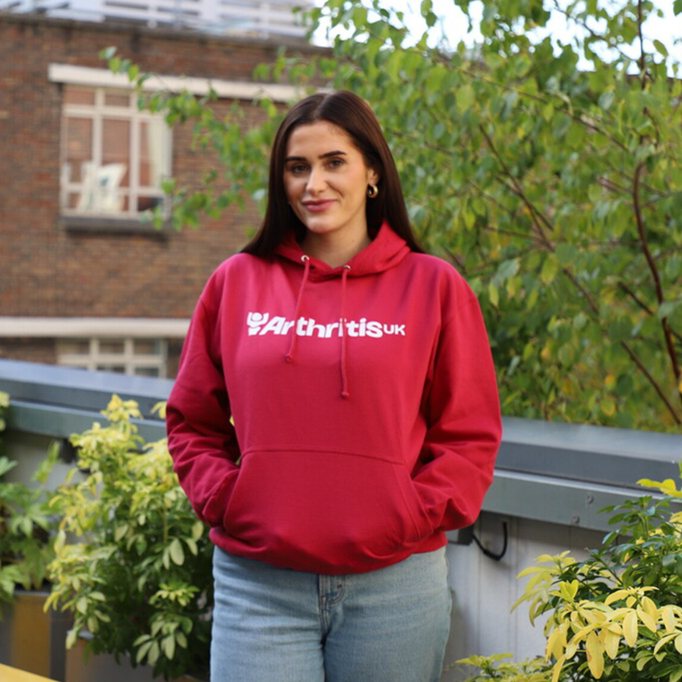 Person wearing a raspberryhoodie with 'Arthritis UK' text on a rooftop with plants and buildings in the background.