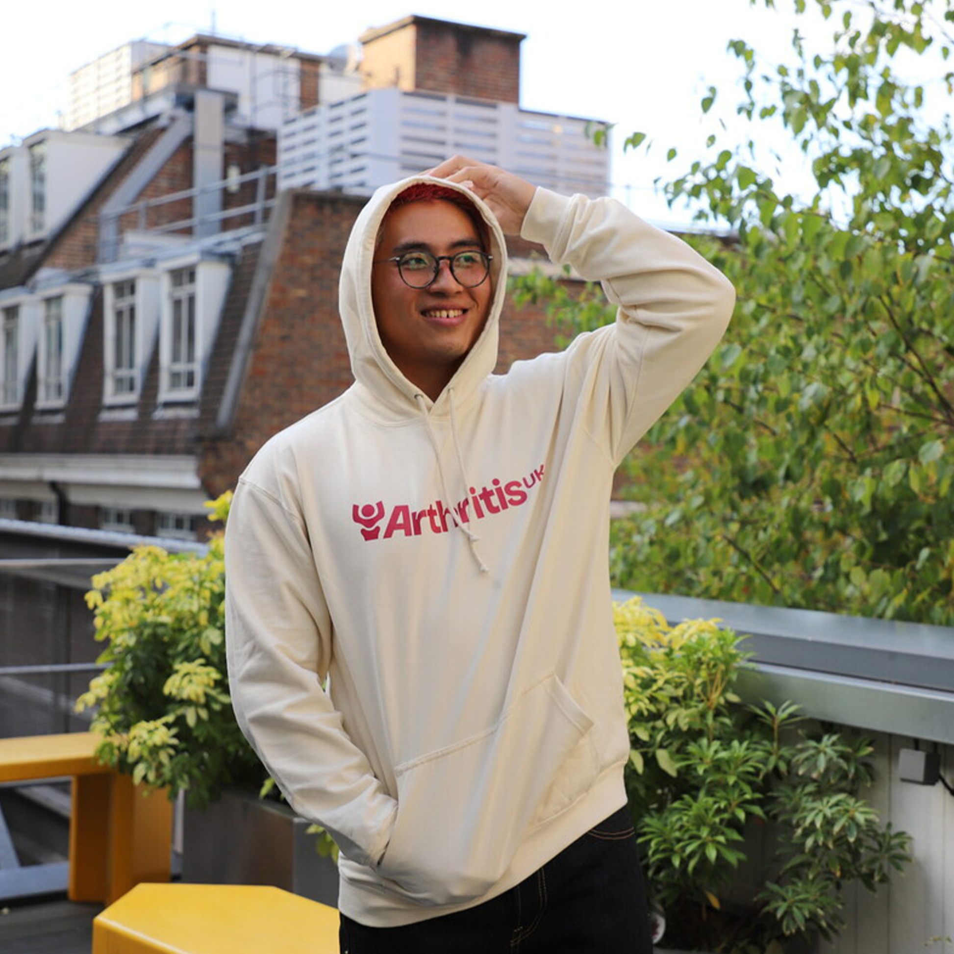 Person wearing a white hoodie with 'Arthritis UK' logo, on a rooftop with plants and buildings in the background
