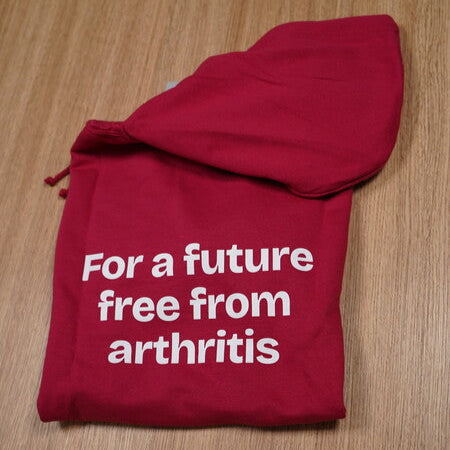 Raspberry hoodie folded up with white text 'For a future free from arthritis' on a wooden surface