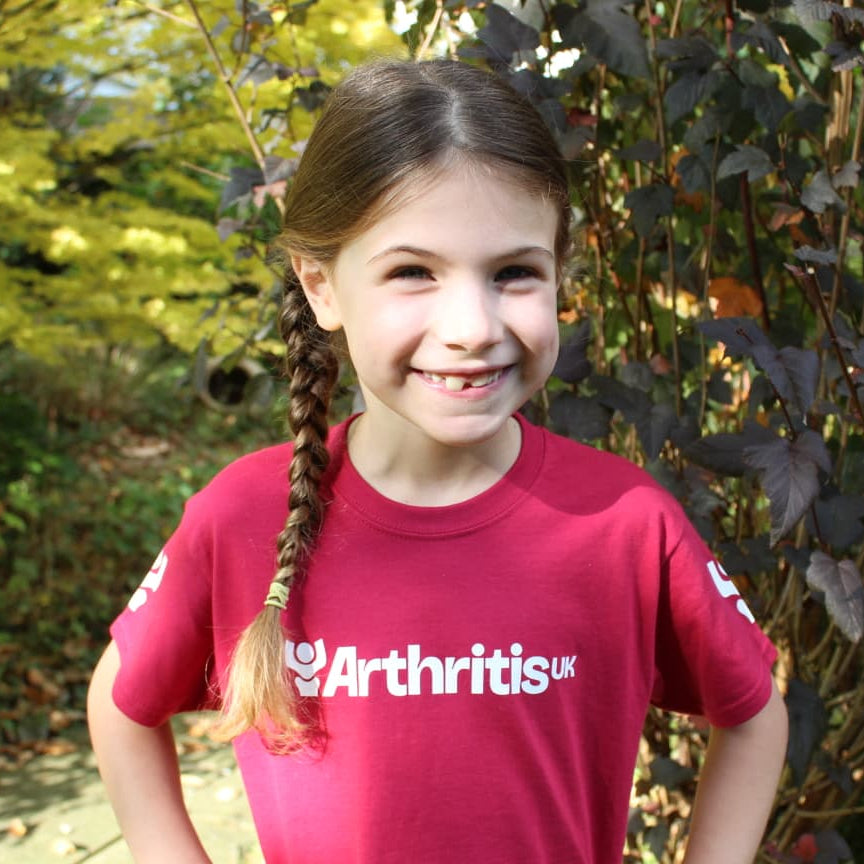 Young girl wearing a raspberry 'Arthritis UK' t-shirt standing outdoors with greenery in the background