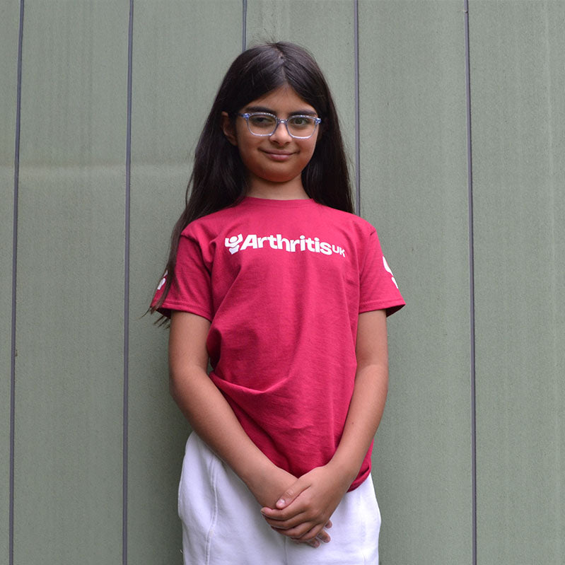 Young girl wearing a raspberry 'Arthritis UK' t-shirt standing against a green wall.