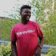 Man wearing a raspberry Arthritis UK t-shirt standing outdoors with greenery in the background