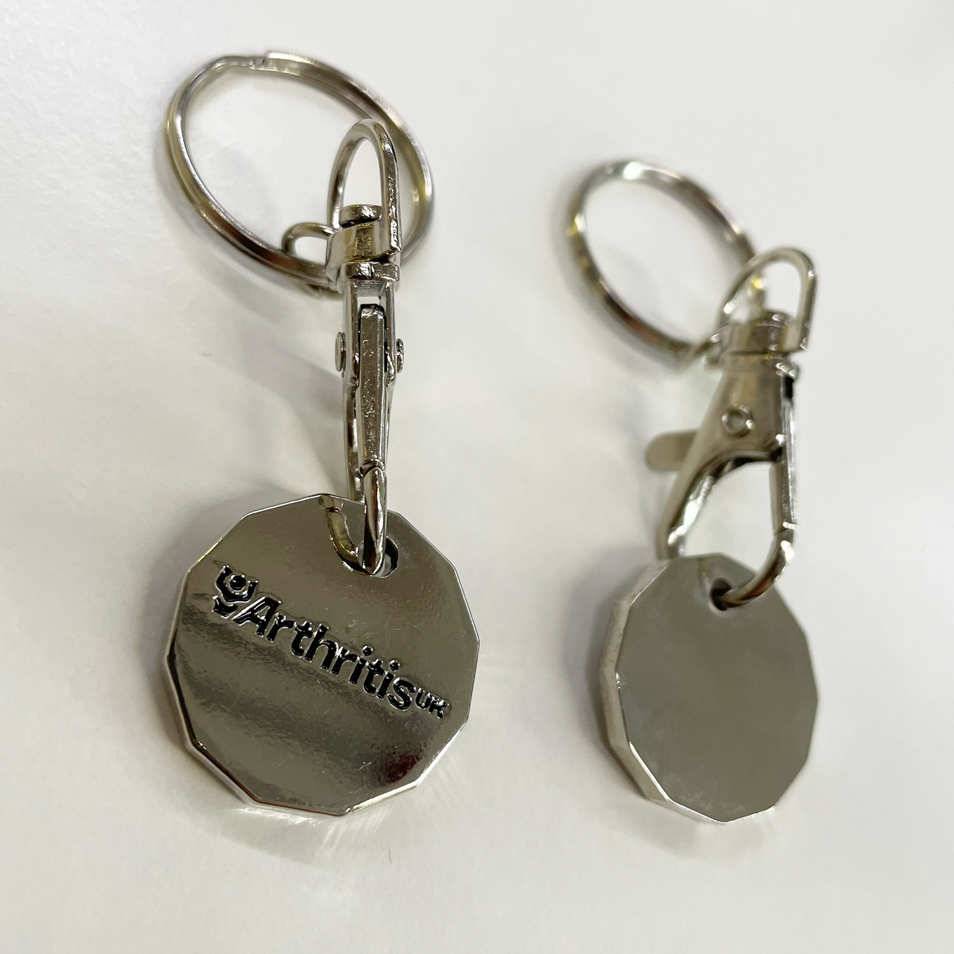 Two silver trolley coin keyrings, silver with the Arthritis UK logo in black.