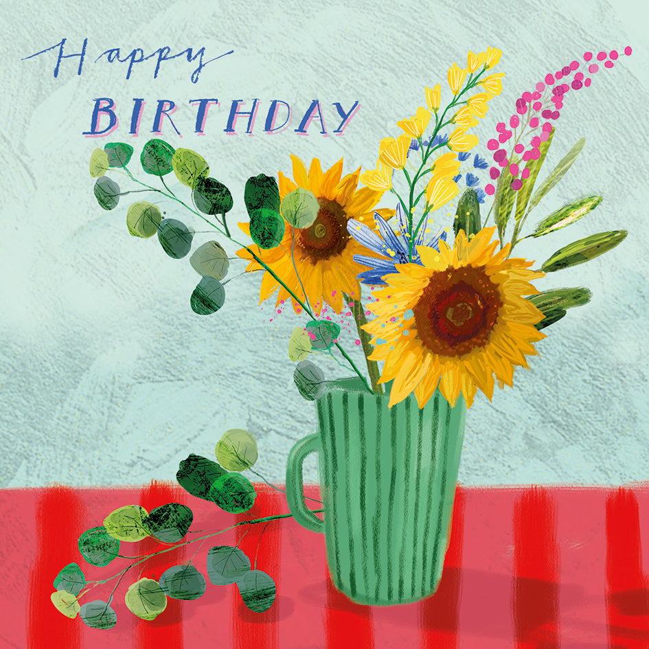 A colorful birthday card illustration featuring a green mug filled with sunflowers, pink blossoms, and green leaves on a red table against a textured light blue background, with the words 'Happy Birthday' in blue and purple at the top.