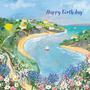 A birthday card with a coastal landscape illustration, including a beach, hills, and a village by the sea. The card has 'Happy Birthday' text.