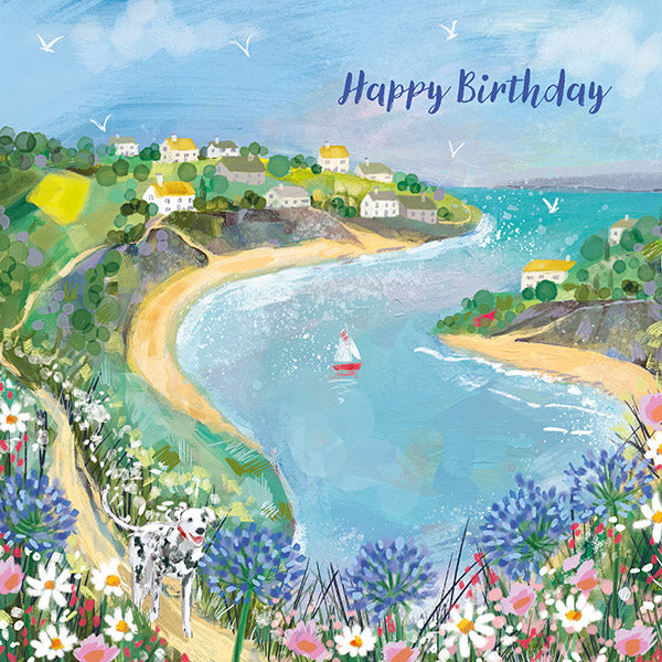 A birthday card with a coastal landscape illustration, including a beach, hills, and a village by the sea. The card has 'Happy Birthday' text.