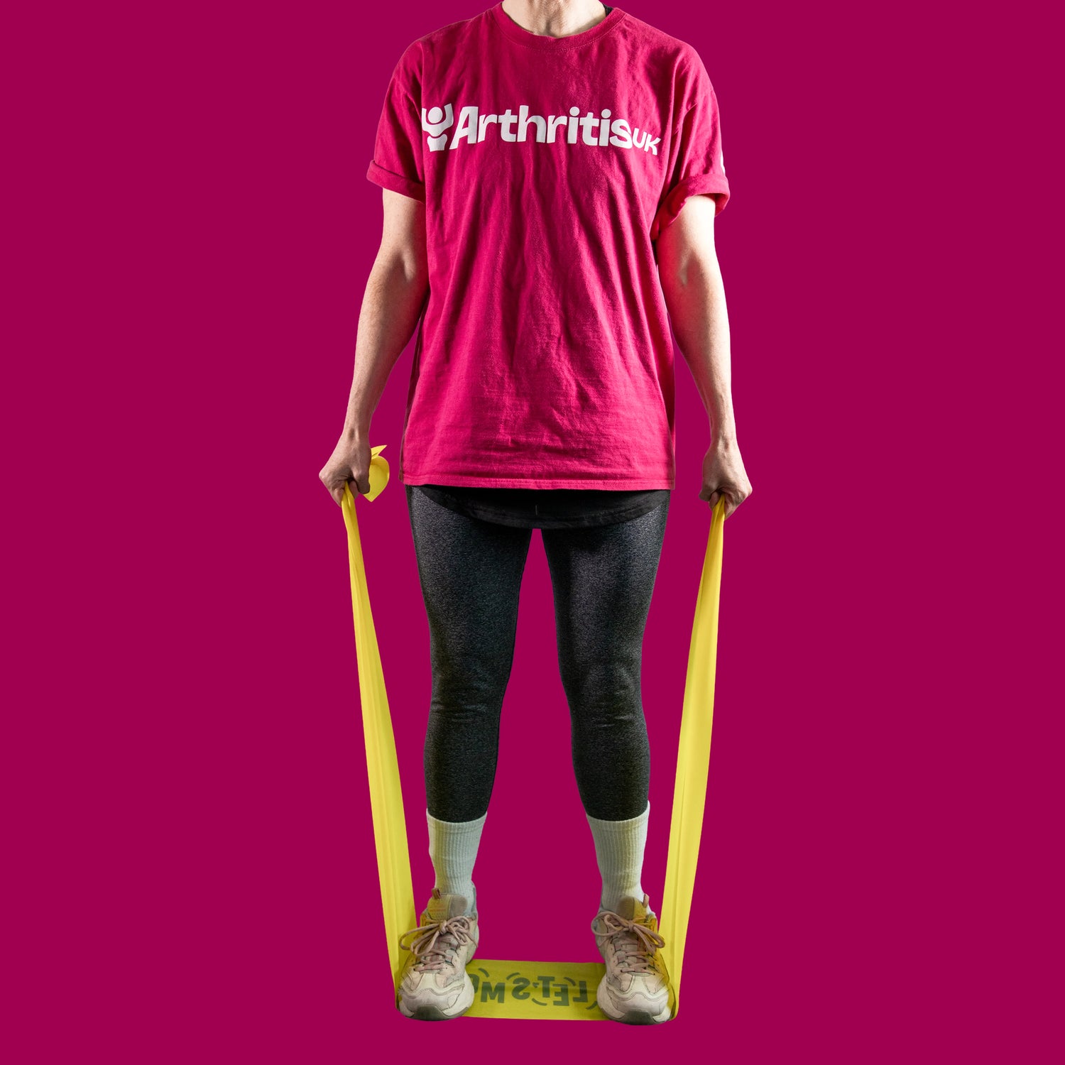 A person standing wearing a bright pink t-shirt with 'Arthritis UK' printed in white across the chest, black leggings, white socks, and beige sneakers. They are holding a yellow resistance band around their feet.