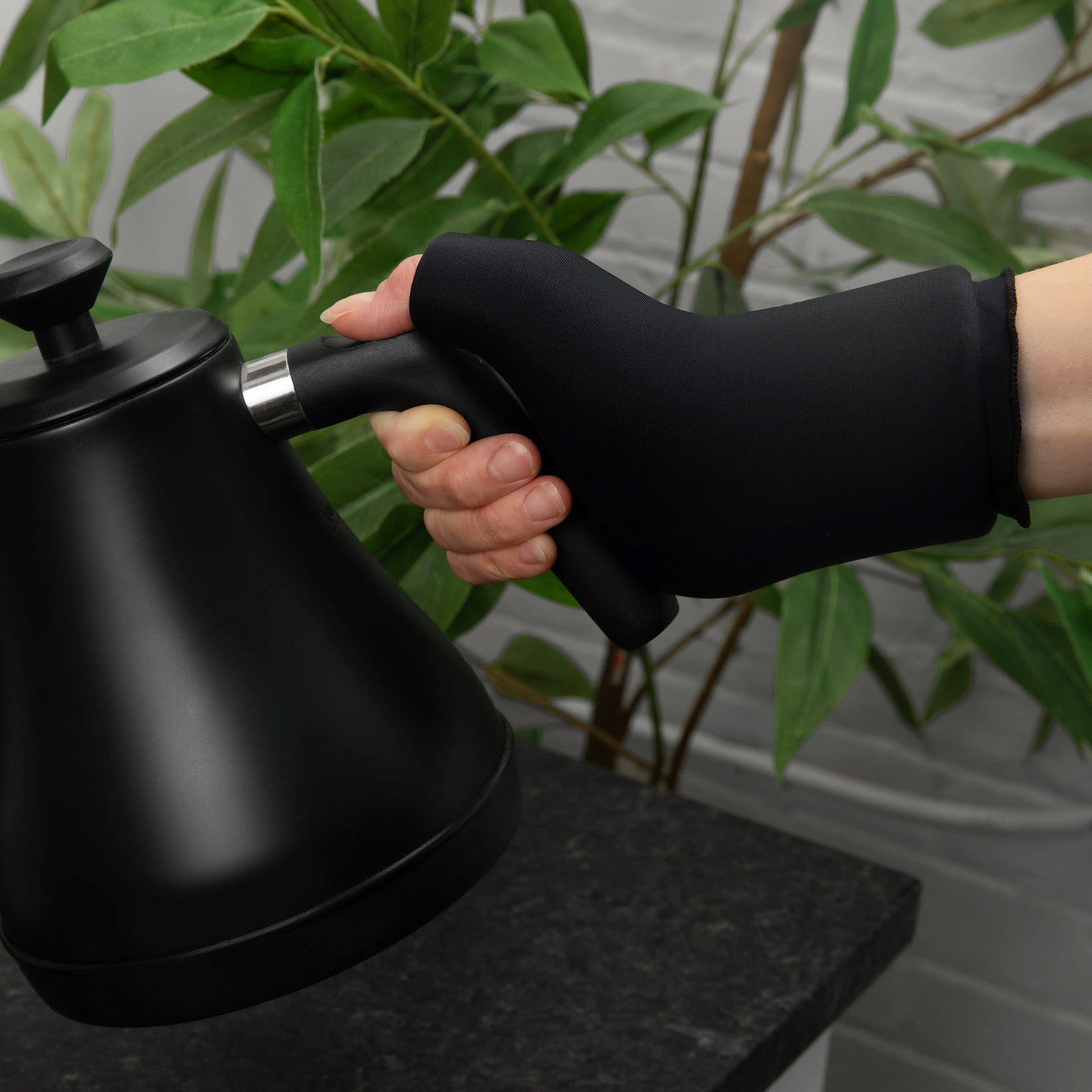 Person holding a black kettle wearing the Grace and Able cooling compression sleeve for thumb and wrist pain with a plant in the background