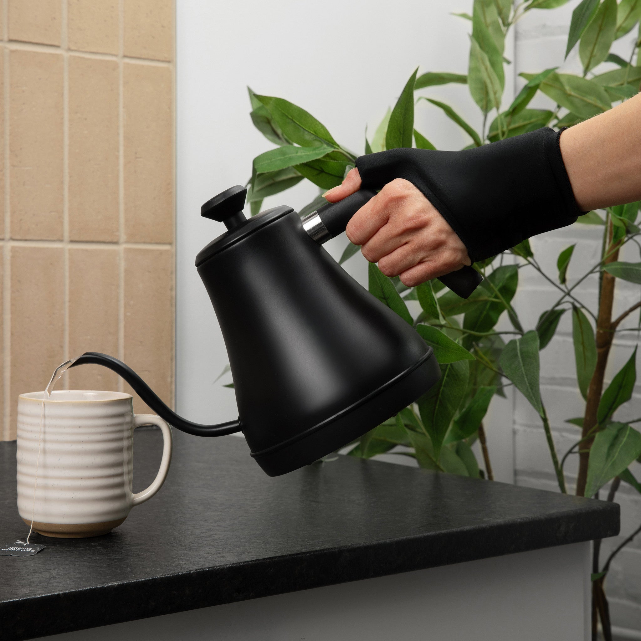 Person wearing the Grace and Able cooling compression sleeve for thumb and wrist pain in black pouring tea from a black kettle into a white mug on a kitchen counter with plants in the background.