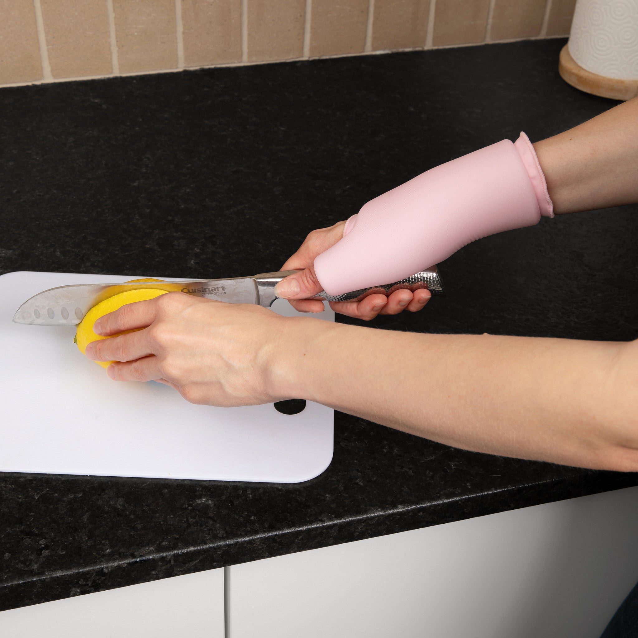 Person using a knife with a yellow handle on a cutting board, wearing a pink Grace and Able Cooling Compression Sleeve For Thumb & Wrist Pain.
