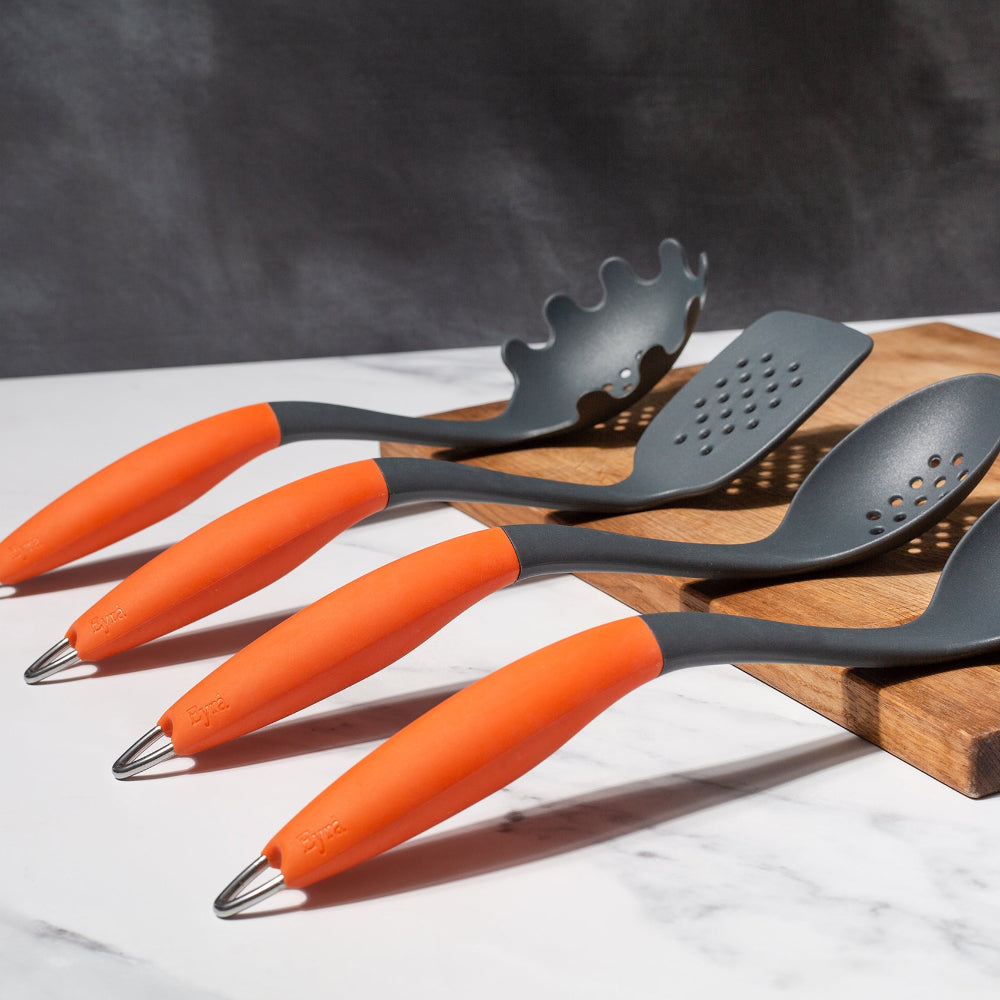 A set of Eyra ergonomic kitchen utensils with orange handles and silver-coloured heads, including a spoon, slotted spoon, spatula, and pasta grabber, displayed on a wooden surface.