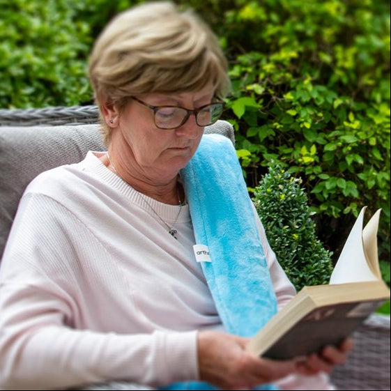 A woman with glasses sitting outdoors, with a long hot water bottle wrapped around her while reading a book. 